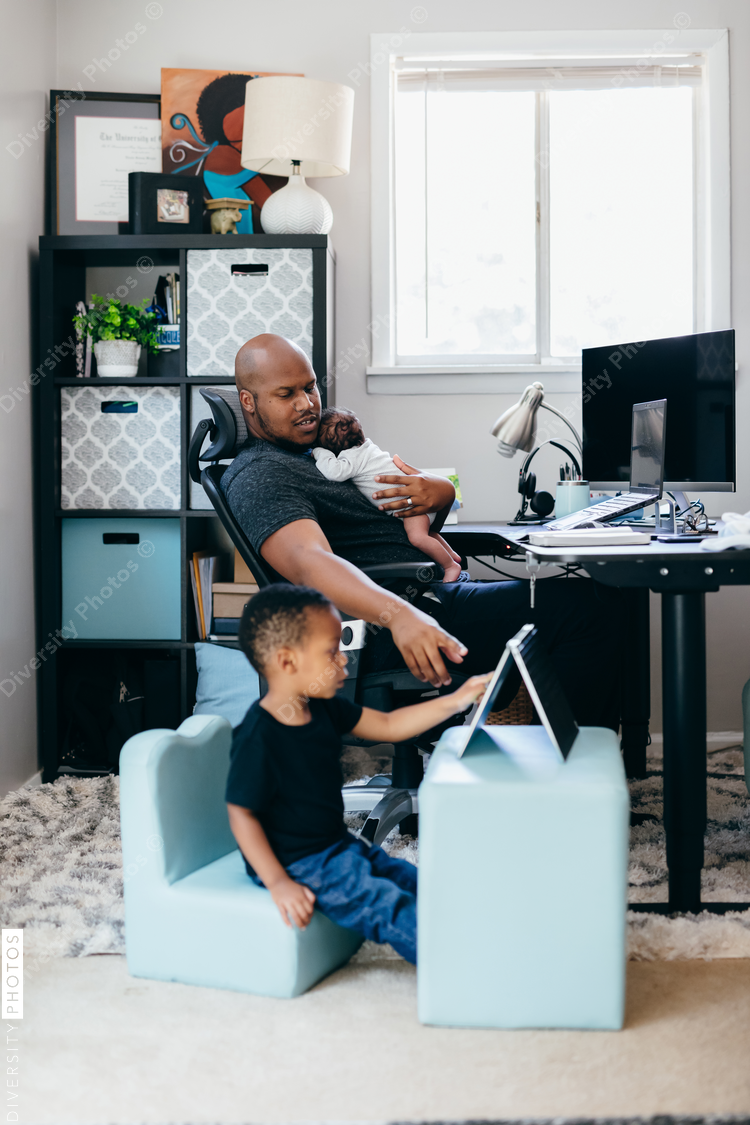 Black dad with newborn working from home helping son with schoolwork