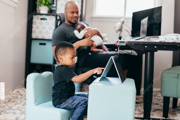 Black dad with newborn working from home helping son with schoolwork