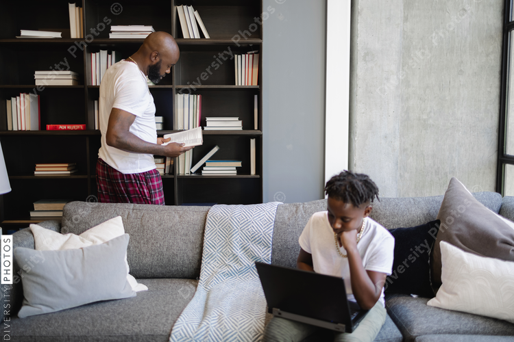 Black Boy using laptop on sofa in living room, father at bookshelf in background