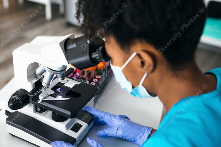 Black medical technologist in mask in laboratory looking through microscope at blood samples