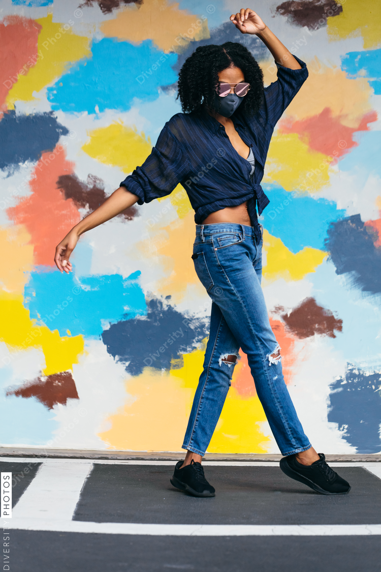 Black woman with natural hair wearing mask and sunglasses dancing in front of mural
