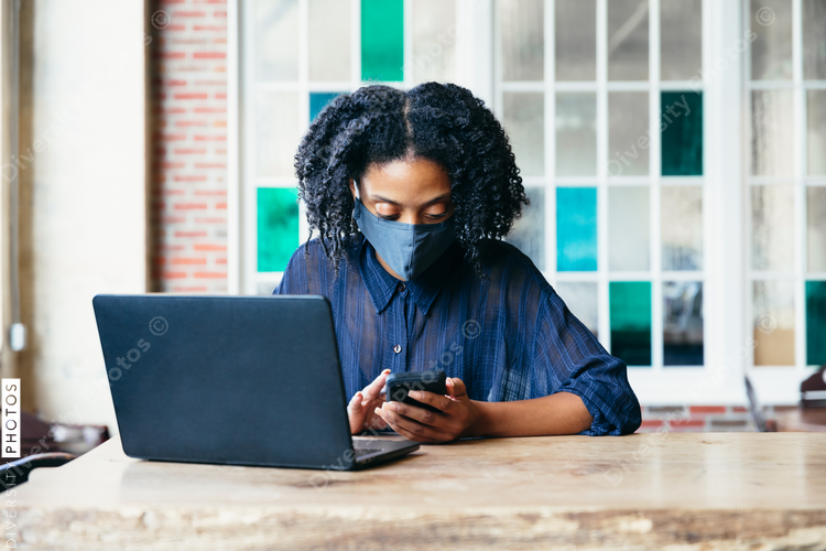 Black woman working at cafe on laptop and phone with face mask during covid-19