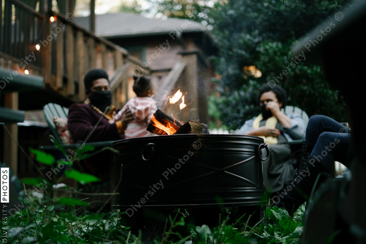 Black friends sitting around firepit safely wearing mask in backyard