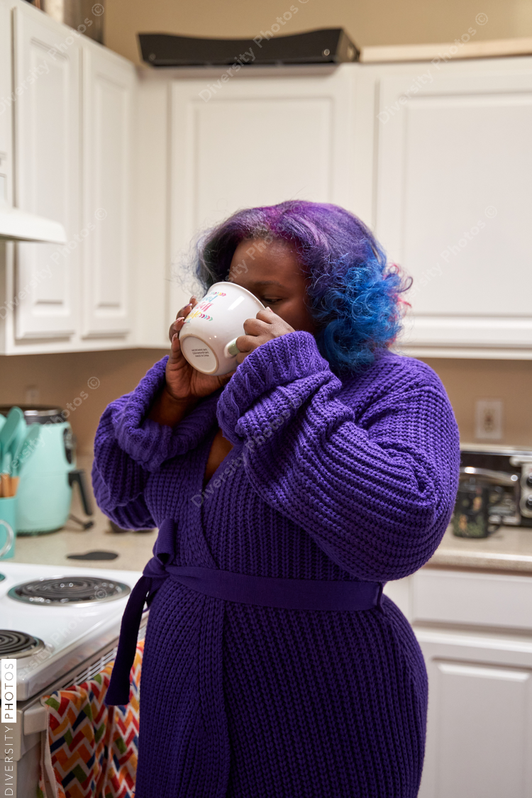 Black woman in robe, drinking morning coffee in kitchen, colorful hair