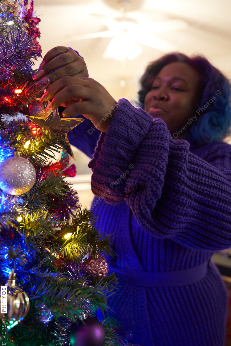 Black woman decorating Christmas tree at home, comfortable in robe