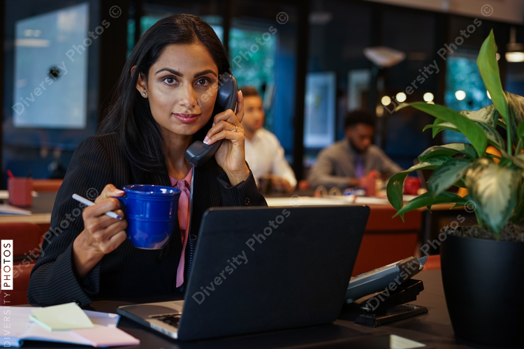 Businesswoman drinking coffee while talking on telephone in office