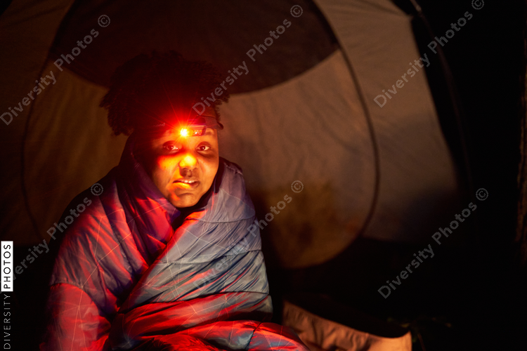 Woman sits around campfire at night wrapped in sleeping bag
