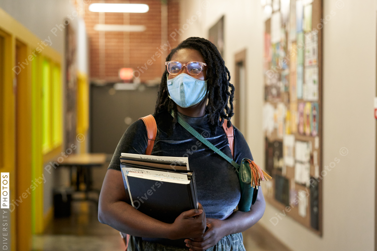 Black female college student walking down hallway with school books, wearing a face mask