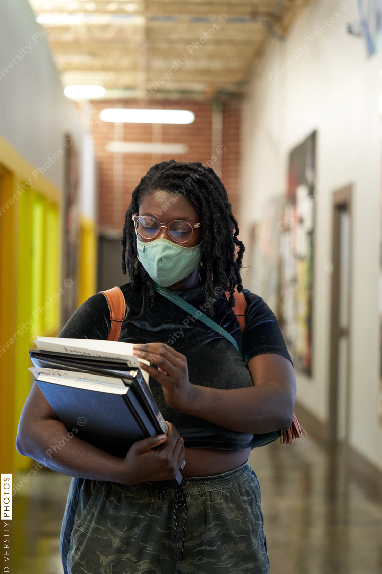 Black female college student walking down hallway with school books, wearing a face mask