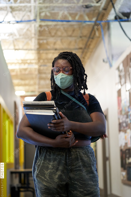 Black female college student walking down hallway with school books, wearing a face mask