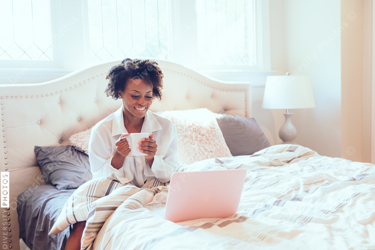 Woman sitting on bed holding coffee cup and looking at laptop