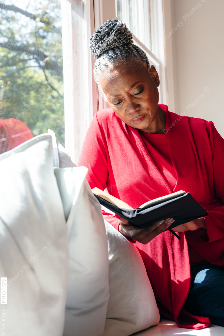 Senior Black woman with grey hair sitting by window and reading book