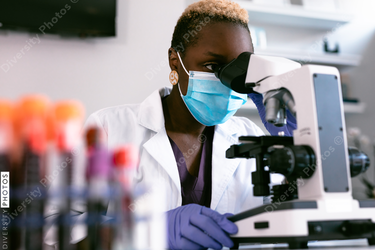 Black female scientist in mask in laboratory looking through microscope