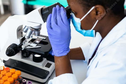 Black medical technician in mask in laboratory looking through microscope