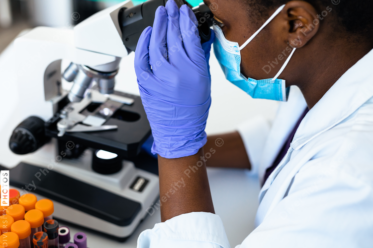 Black medical technician in mask in laboratory looking through microscope