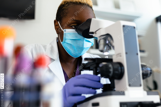 Black medical technologist in mask in laboratory looking through microscope at blood samples
