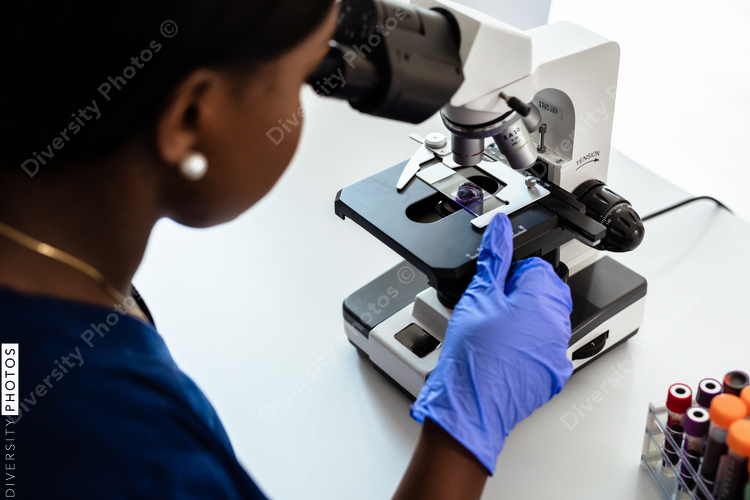 Black female medical professional in laboratory looking through microscope at blood samples