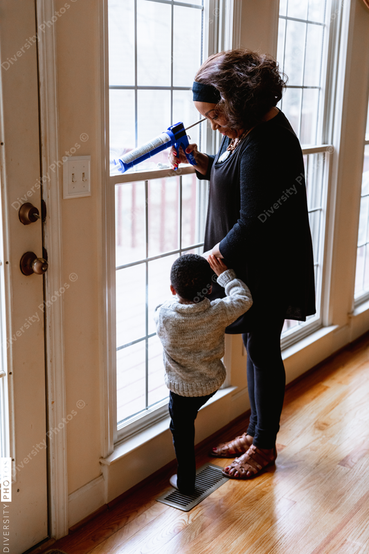 Black grandmother woman weather sealing, caulking windows for winter with toddler grandson