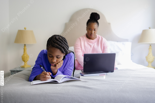 Black Teenage girl lying on bed and writing in notebook, mother using laptop in background