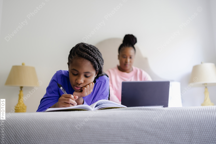 Teenage girl lying on bed and writing in notebook, mother using laptop in background
