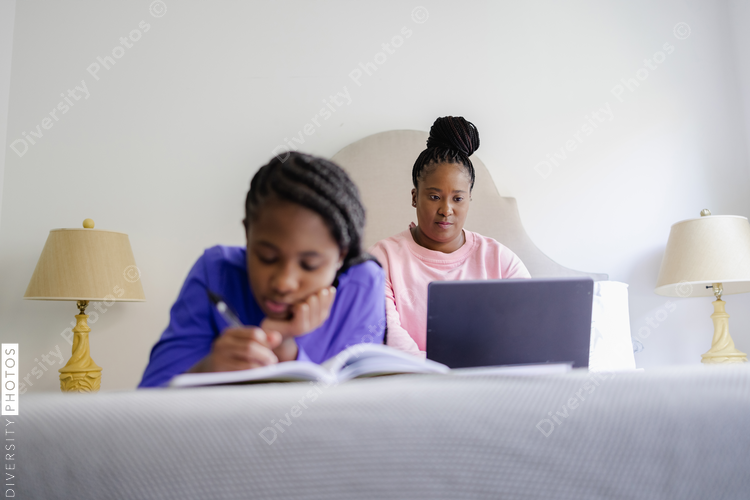 Teenage girl lying on bed and writing in notebook, mother using laptop in background