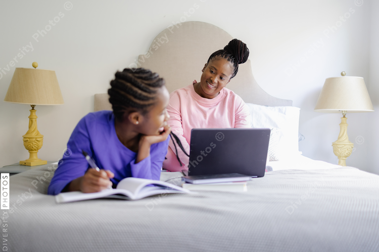 Black Teenage girl lying on bed and writing in notebook, smiling to mother using laptop in background