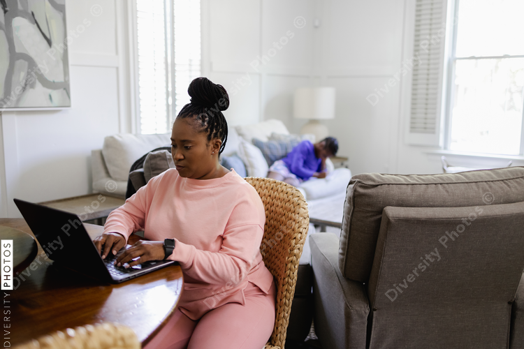Black Woman using laptop at table in living room, teenage daughter lying on sofa in background