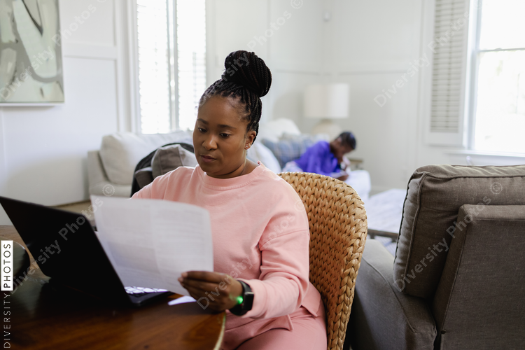 Black Woman using laptop and reading document at table in living room, teenage daughter lying on sofa in background