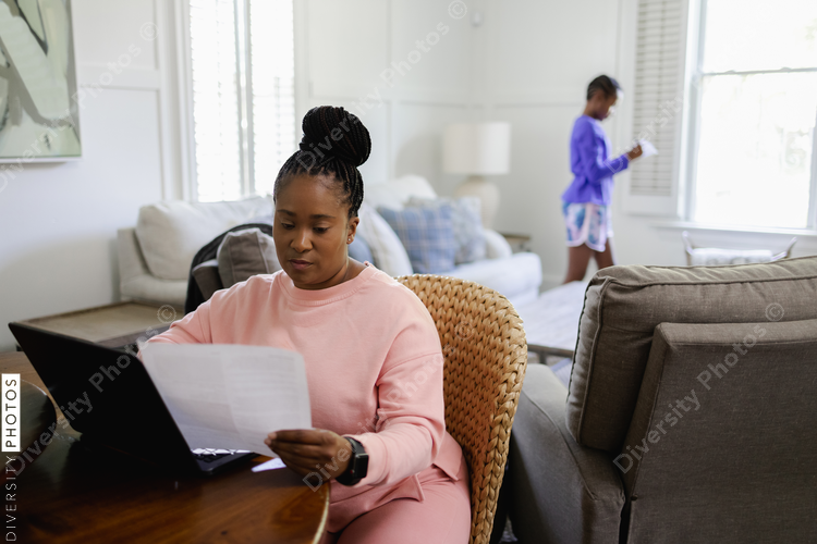 Woman using laptop and reading document at table in living room, teenage daughter in background