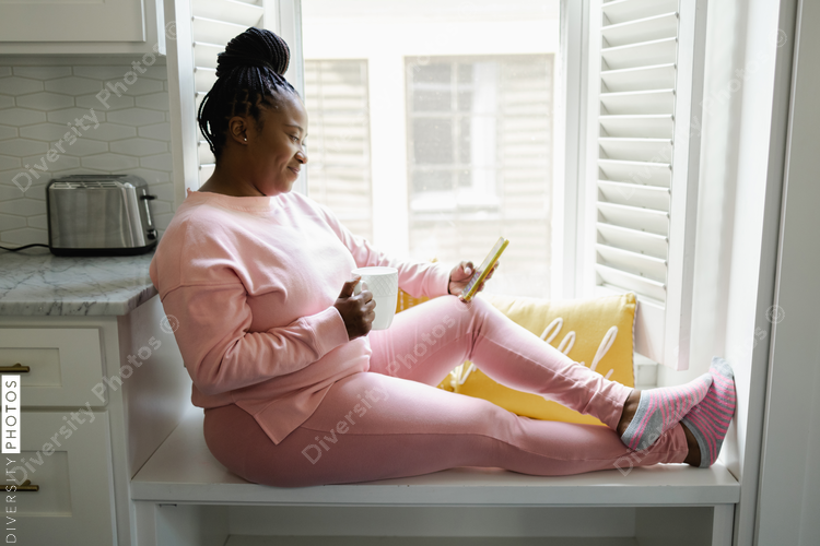 Smiling woman with braided hair sitting by window with smart phone and mug, African American