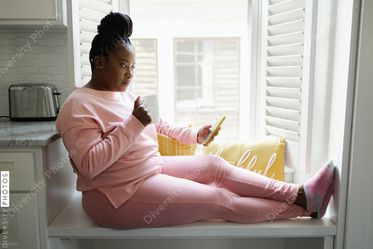 African American Woman with braided hair sitting by window with smart phone and mug