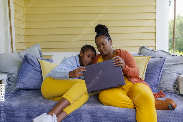 Black Mother and teenage daughter sitting on porch swing, looking at laptop