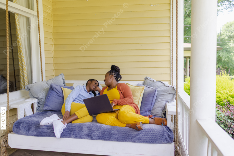 Smiling mother and teenage daughter sitting on porch swing with laptop