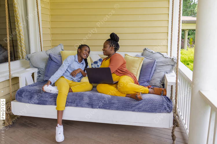 Smiling mother and teenage daughter sitting on porch swing with laptop