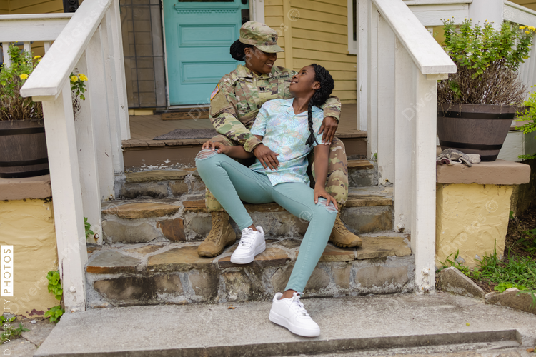 Happy military woman in camouflage uniform sitting with teenage daughter on steps in front of house