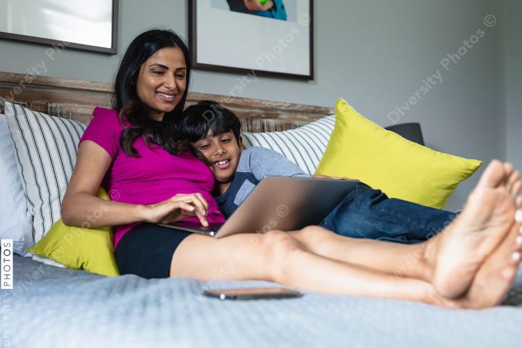Indian mother and son snuggles in bed while mom is working on laptop