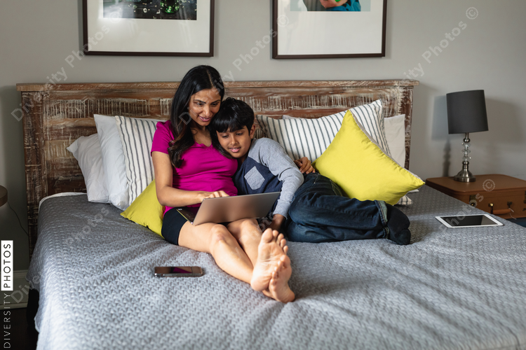 Indian mother and son snuggles in bed while mom is working on laptop