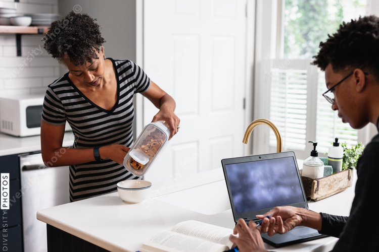 Black mother preparing breakfast cereal for son studying on laptop