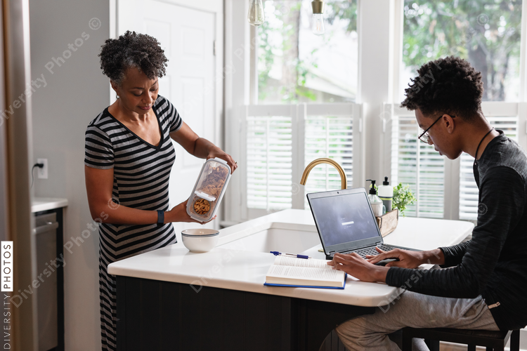 Black mother preparing breakfast cereal for son studying on laptop