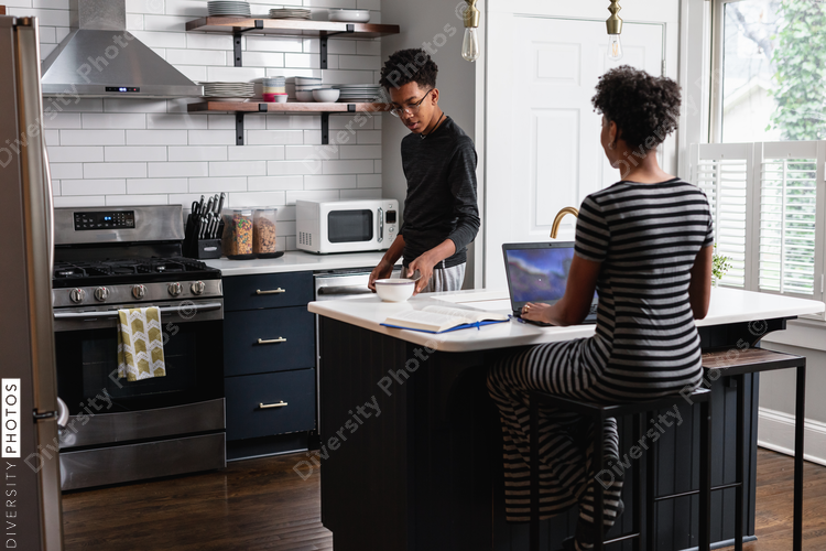 Black son prepares cereal breakfast food while mom is working on laptop