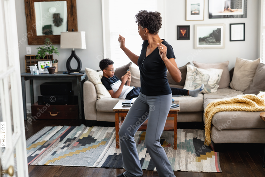 Black mom enjoys music, dancing in family room, son in background, weekend morning