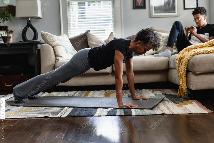 Black mom working out in family room, teenage son on phone in background, planks