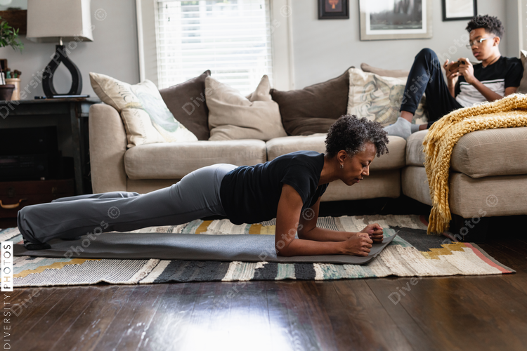 Black mom working out in family room, teenage son on phone in background, planks