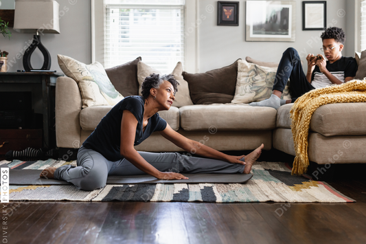 Black mom stretches in family room, teenage son on phone in background, fitness, health and wellness