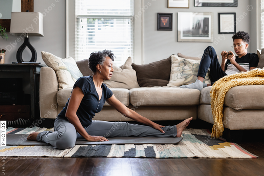 Black mom stretches in family room, teenage son on phone in background, fitness, health and wellness