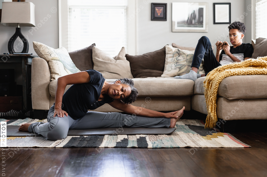 Black mom stretches in family room, teenage son on phone in background, fitness, health and wellness