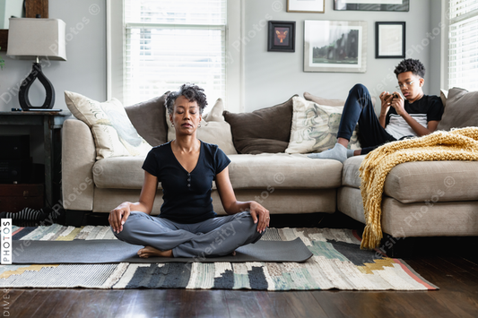 Black mom meditates in family room, teenage son on phone in background, peaceful health  and wellness