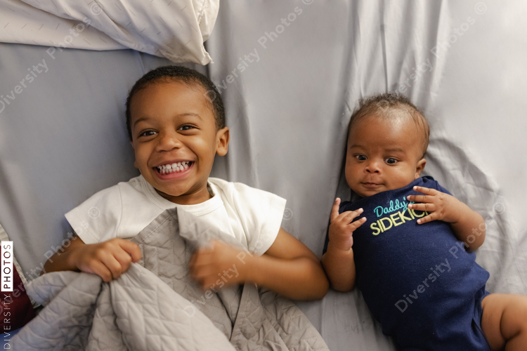 Two African American brothers, 2 yrs old and newborn, laying on bed together, happy