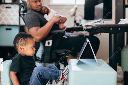 Black dad with newborn working from home helping son with schoolwork 