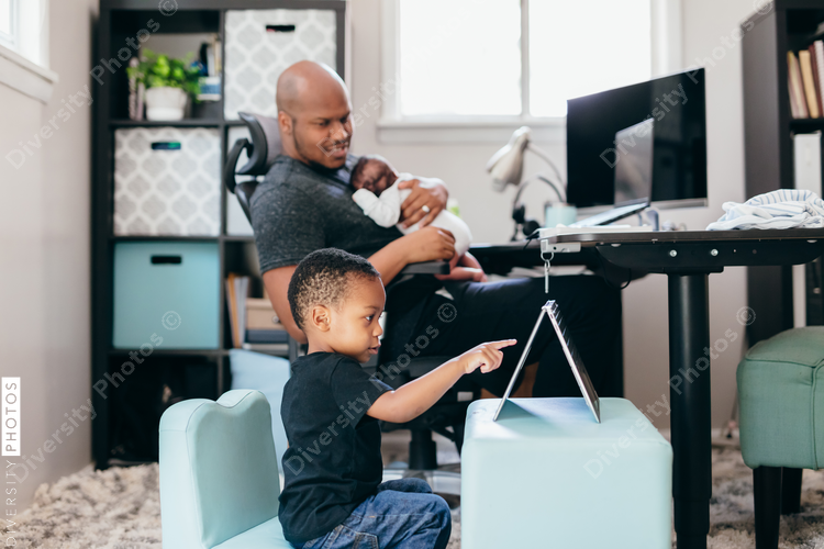 Black dad with newborn working from home helping son with schoolwork 